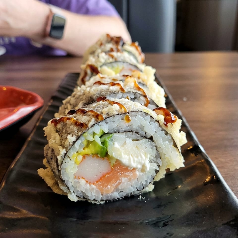 Close-up of a sushi roll topped with crispy coating and sauce on a black rectangular plate, with pickled ginger and diner in background.