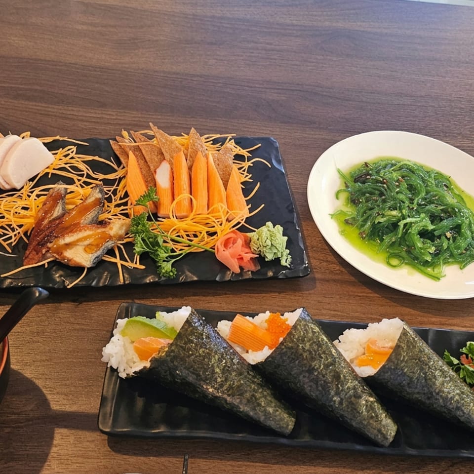 Japanese meal featuring sashimi, hand rolls, seaweed salad, miso soup, and garnishes on a wooden restaurant table.